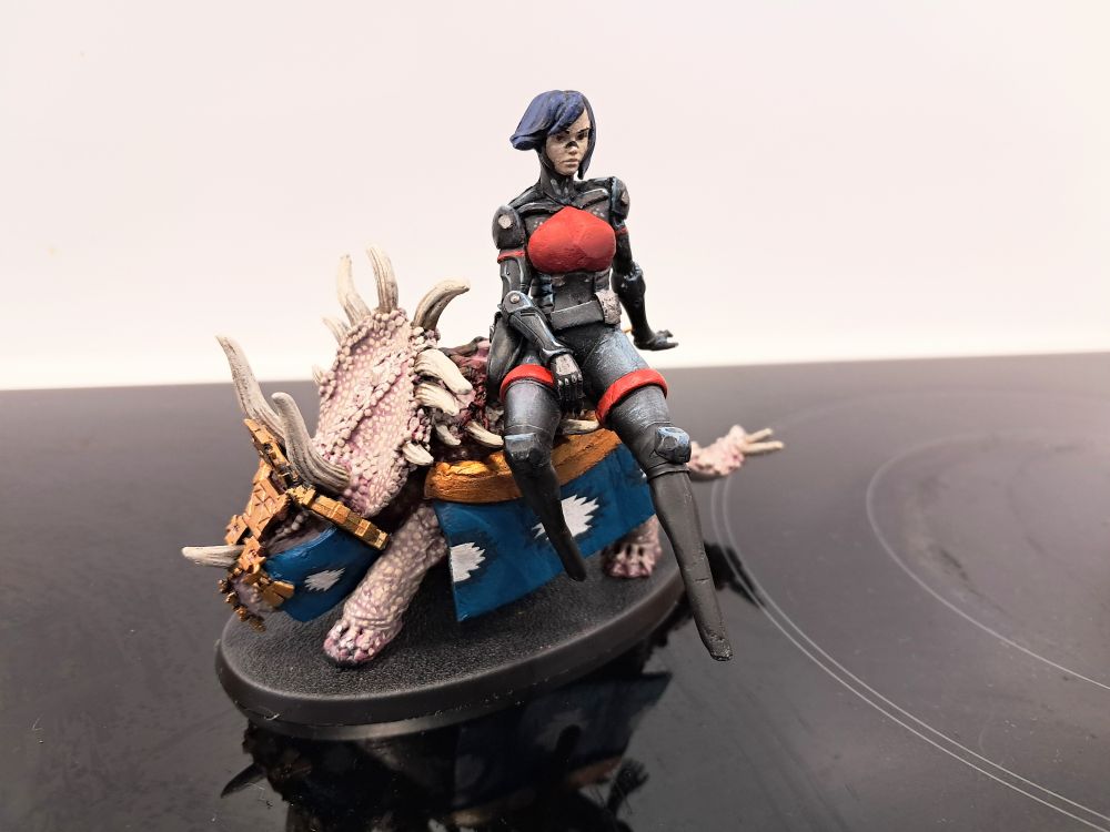 Photograph depicting a "75mm scale" kitbashed Elster Mini from Signalis, sat on top of a classic metal stegadon from Warhammer fantasy that I happened to be working on at the same time. She doesn't seem nearly as thrilled as I would be to be sitting on a pint-sized dinosaur, but I suppose she misses her wide so I understand 