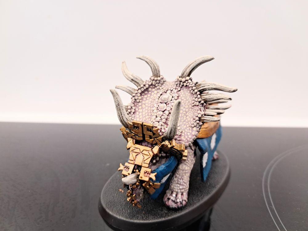 Photograph depicting a classic metal stegadon, with the howdah removed and kitbashed golden armour on the head and fabric skirting on the sides and face
The stegadon has been painted to look like it is both albino and extremely old, with lots of decorations to venerate it in its old age.
This photo shows the animal from the front