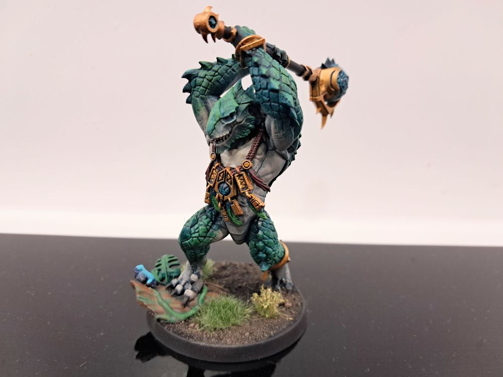 Photograph of the lead kroxigor. They're striding through jungle vegetation and are themselves a sea green/teal with pale bellies. Their metallic ornamentation and weapon details are a vibrant golden material, and their celestite weapons are a translucent turquoise crystal.
They bear a drakespite mace held high over their head, and a poison dart frog rests on the log they're stepping on.