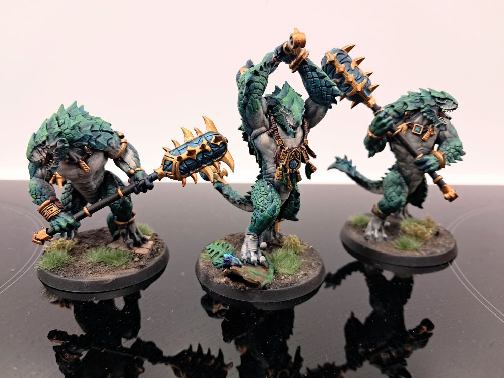 Photograph of three fully painted kroxigor. They're striding through jungle vegetation and are themselves a sea green/teal with pale bellies. Their metallic ornamentation and weapon details are a vibrant golden material, and their celestite weapons are a translucent turquoise crystal.