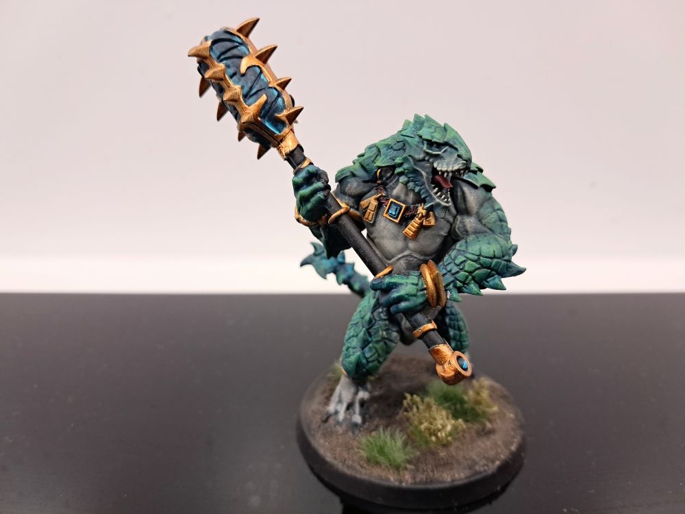 Photograph of a fully painted kroxigor. They're striding through jungle vegetation and are themselves a sea green/teal with pale bellies. Their metallic ornamentation and weapon details are a vibrant golden material, and their celestite weapons are a translucent turquoise crystal.
They are hefting their maul and roaring.