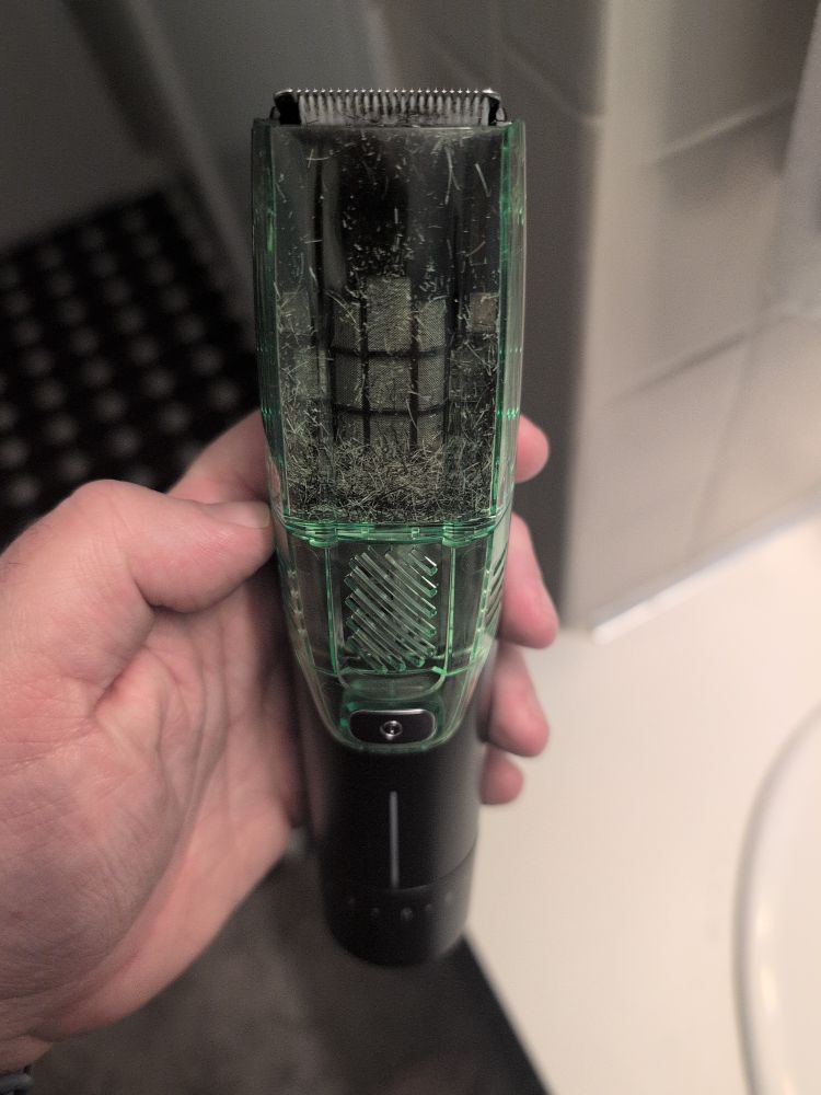 A beard trimmer with a bright green vacuum bin ~10% full of brown and white hair