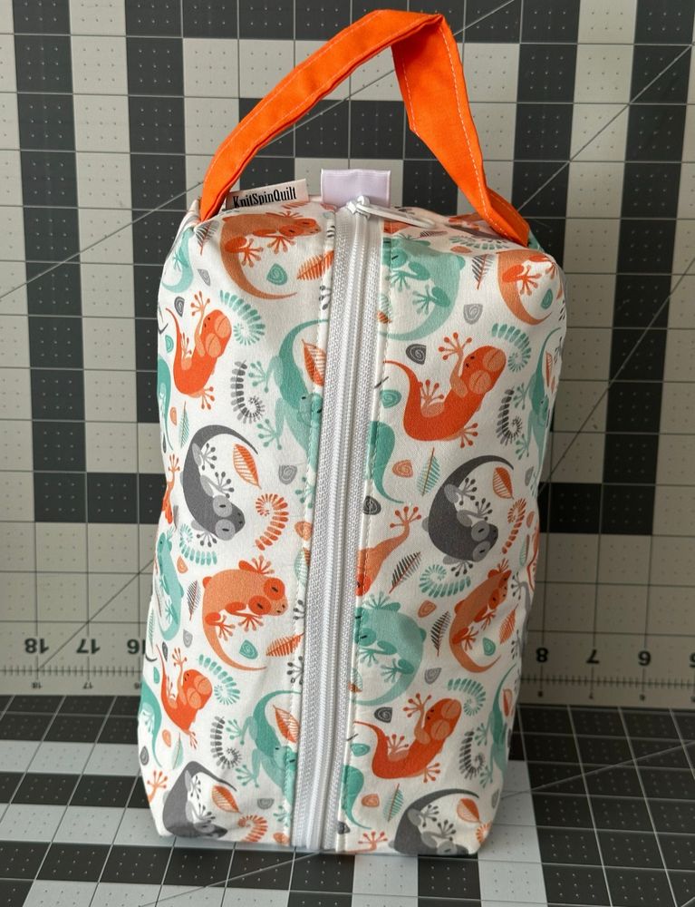 A box bag standing on end. The fabric is a white background with tessellated geckos on it in orange, green, and grey. The bag’s handle is orange. 
