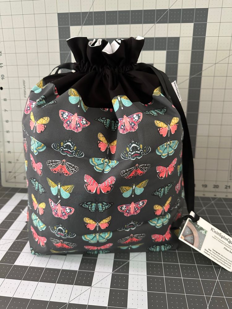 A large drawstring bag with a black top sitting on a gridded cutting mat. The main fabric is a grey solid background on which are arrayed columns of colorful moths and butterflies. 
