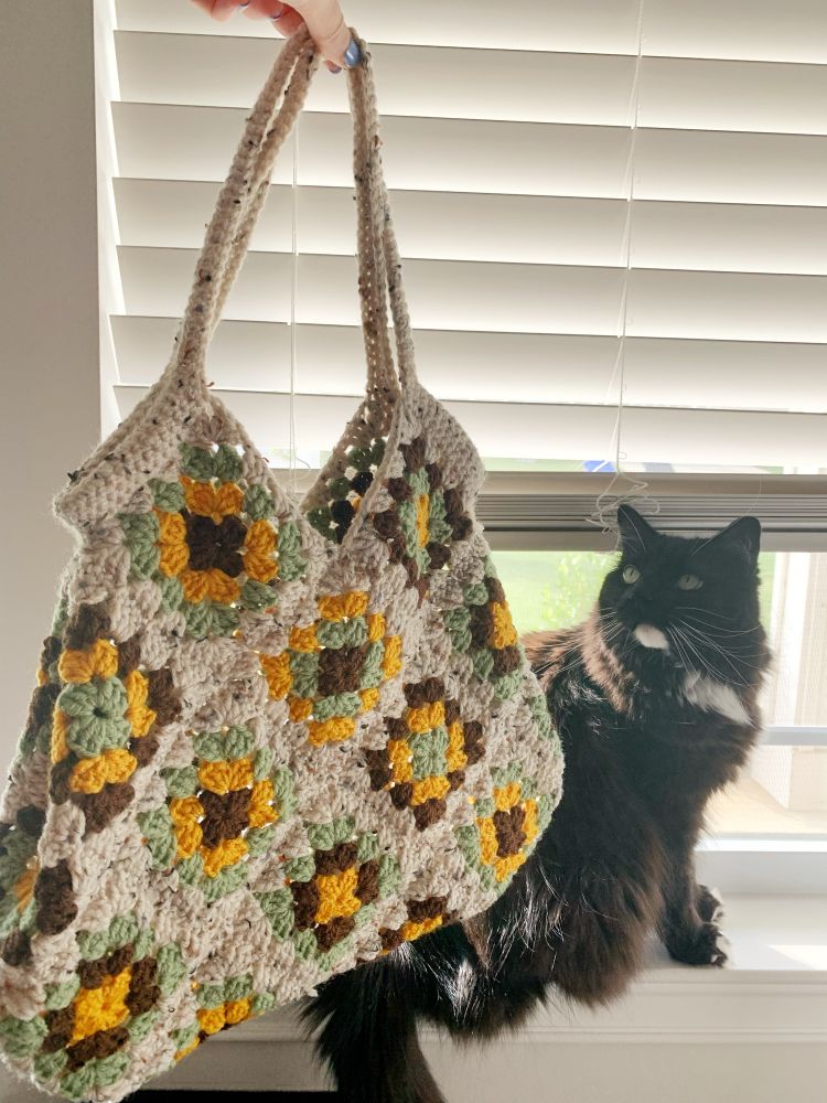 Cherry crocheted bag with granny squares in brown, yellow, and green. A speckled off-white thread brings it all together. But the REAL star of the show is a handsome tuxedo cat curiously looking up at the bag while he soaks up sun on a windowsill