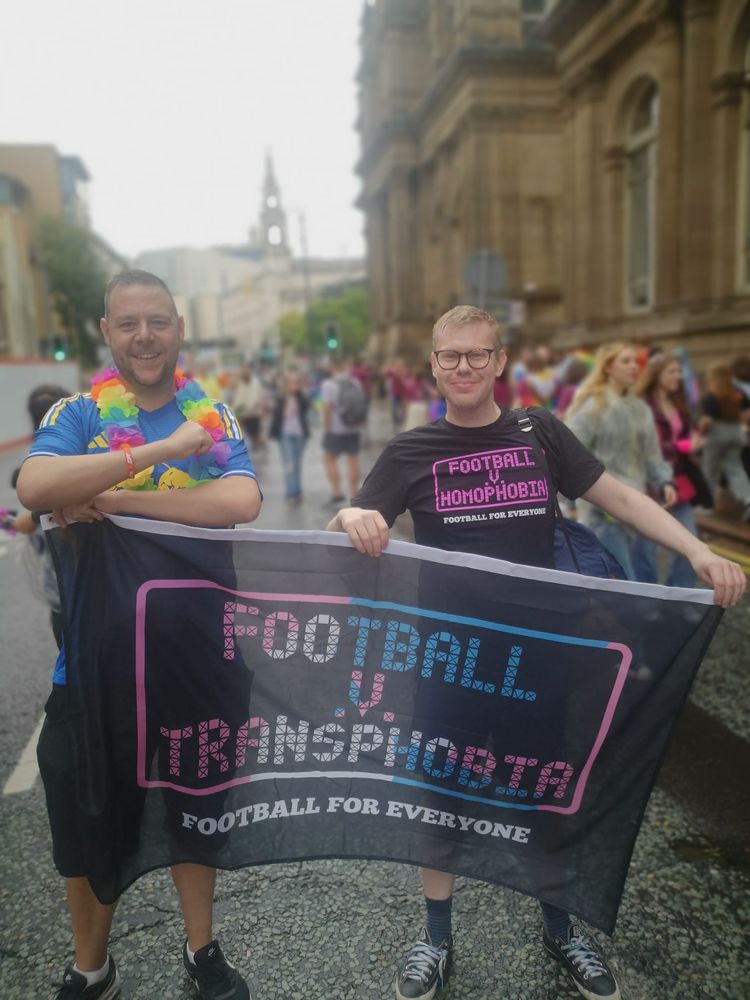 A photo of Rainbow Owls and Marching Out Together's chairs holding the Football v Transphobia flag at Leeds Pride, with the former wearing a Football v Homophobia T-shirt and the latter wearing a blue Leeds United away strip.