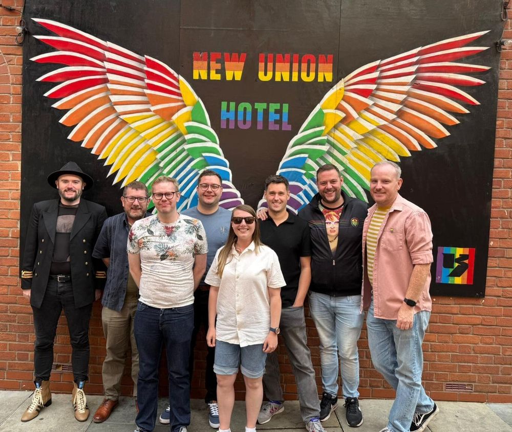 A photo of Regional Alliance members Rainbow Rams, Rainbow Spireites, Proud Stags, Rainbow Blades, Marching Out Together, Proud Forest and Rainbow Owls standing together behind a New Union Hotel mural on Canal Street.