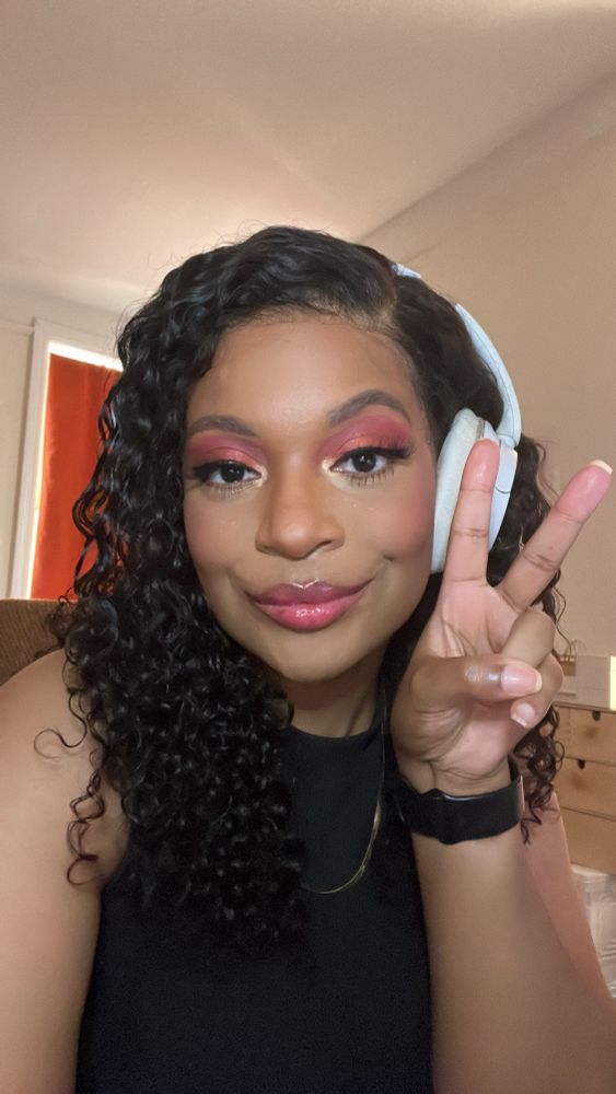 a selfie of Jupiter, a Black non-binary person. They have curly hair that is parted to the side. They’re wearing white headphones, a black shirt, and a gold necklace. They’re wearing pink and red makeup. They are holding up a peace sign. 