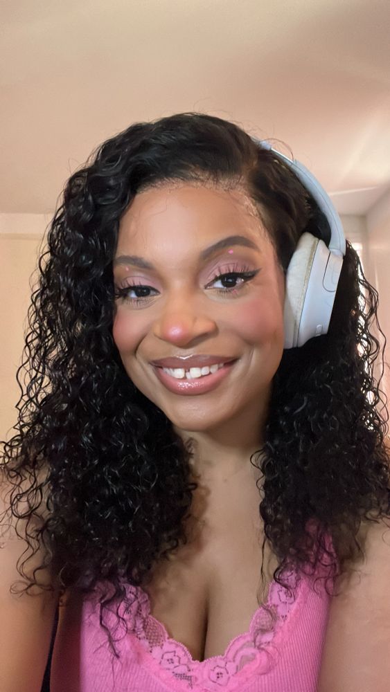 A selfie of Jupiter, a Black non-binary person. They have dark curly hair, and are wearing white headphones and a pink camisole top. They’re smiling at the camera. 