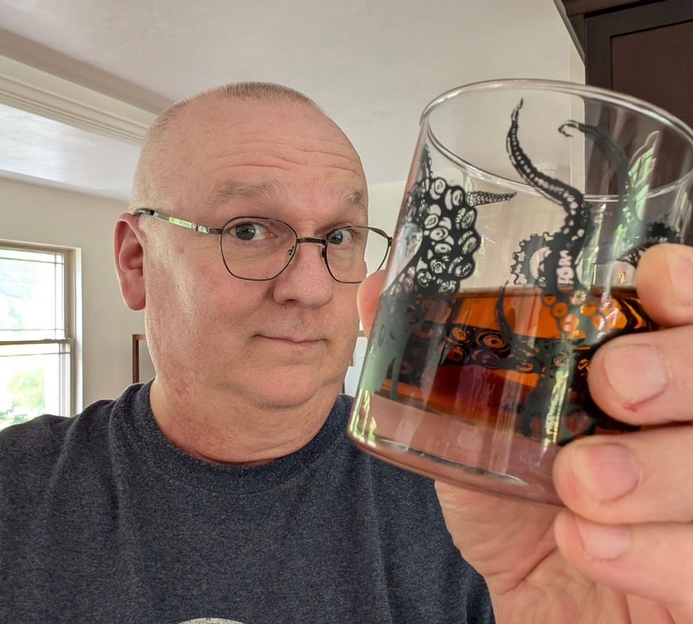 An older, bald'ish man with glasses smiling and holding up a whiskey glass 1/3 full of whiskey. The glass design has tentacle images embossed on it.