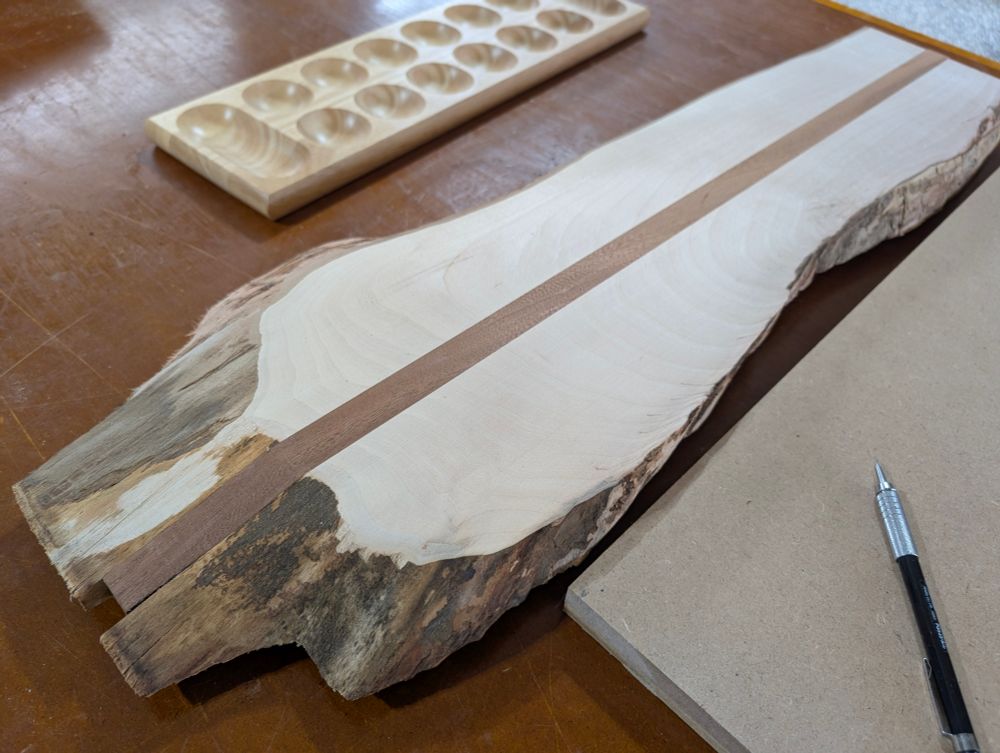 A small, live edge Maple wood slab with a light brown strip of Sapele down the center. A commercially made Mancala board is in the background. 