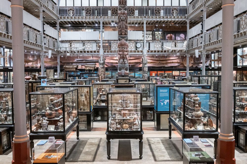 View across museum gallery with densely packed display cases towards wooden crest pole carved with faces.