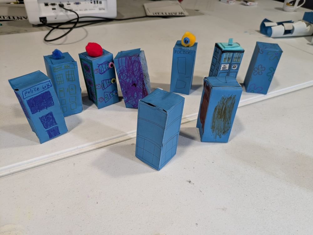 Nine pale blue boxes are clustered together on a table. They represent police boxes, but are designed in different ways using felt tip pens and other items that have been glued on. A police box is often referred to as a Tardis in the UK because of the Tv programme, Dr Who.