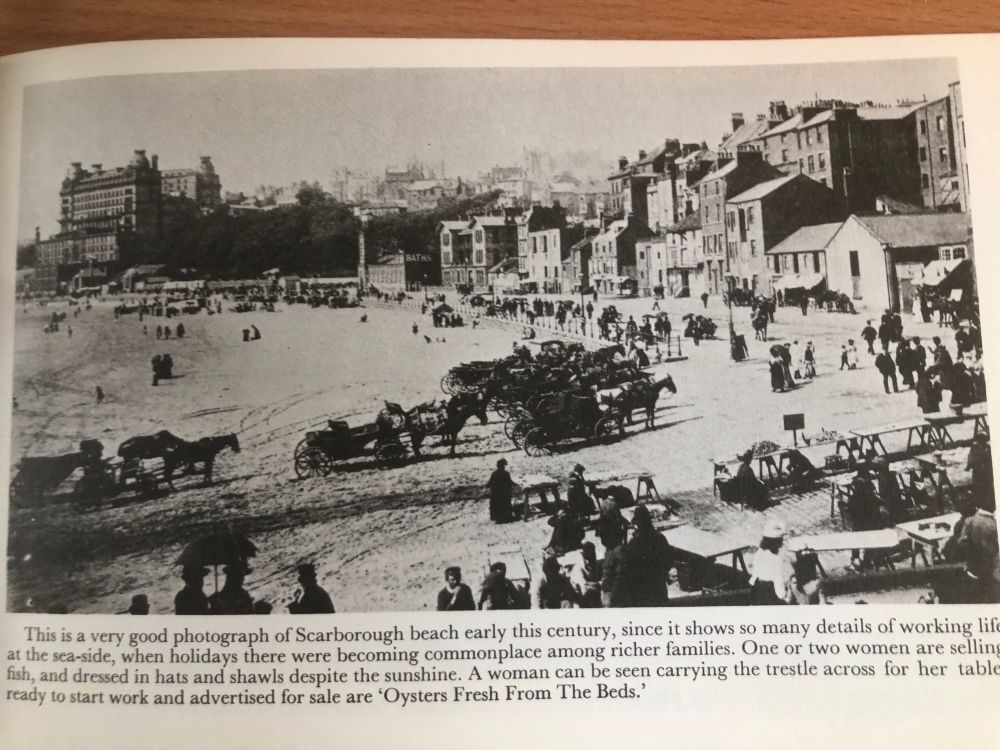 This pic shows a busy beach with holidaymakers, horses be carriages and people selling food. The beach road is lined with buildings and there is a large hotel on the cliff above. The text reads - This is a very good photograph of Scarborough beach early this century, since it shows so many details of working life at the seaside, when holidays were becoming commonplace among richer families. One or two women are selling fish, and dressed in hats and shawls, despite the sunshine. A woman can be seen carry the trestle across for her table ready to start work and advertiser are ‘Oysters fresh from the Beds’.
