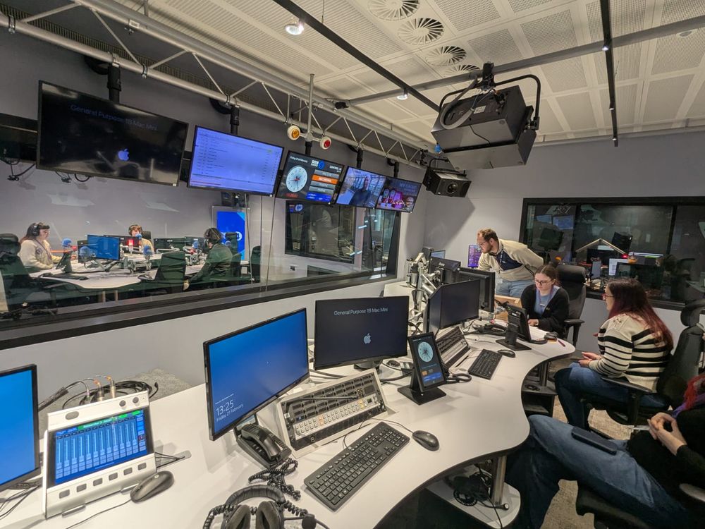 Photo of radio studio with people at mixing desk and others in the studio