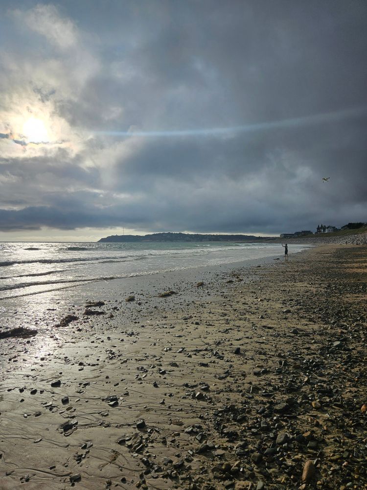 Ein Bild von einem Strand mit Wellen, dicken Wolken und hervorbrechenden Sonnenstrahlen im Gegenlicht. Im Hintergrund eine Landzunge. Am Bildrand ist ein Junge mit einem Drachen zu erkennen.
