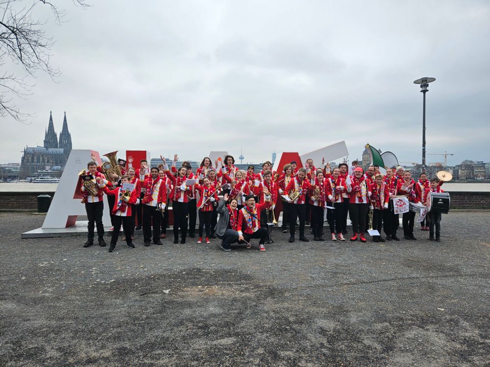 Das Jugendmusikkorps Jumikk posiert vor dem Kölner Alaaf-Zeichen. Sie sind in rot-weiße Uniformjacken gekleidet und tragen schwarze Hosen. Im Hintergrund ist der Kölner Dom zu erkennen.