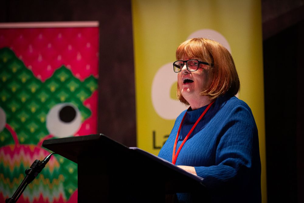 Photograph of Laureate na nÓg, Patricia Forde, standing at a lectern. She has red hair and is wearing a blue dress. She wears glasses and a red lanyard is just visible. In the background there are two pull-ups partially visible, those of the Baboró Festival and Laureate na nÓg. Photo by Anita Murphy.