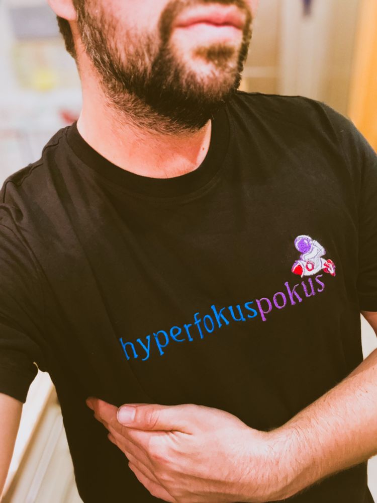 Selfie: Oberkörper und Mundpartie. Ich trage ein schwarzes T-Shirt mit Stickmotiv. Dieses besteht aus dem Schriftzug „hyperfokuspokus“ (wobei hyperfokus in Blau und pokus in Lila gehalten ist), sowie aus einem kleinen Astronauten im Clip-Art Style, der eine Rakete reitet. Dieser ist über dem rechten Ende des Schriftzug positioniert. 