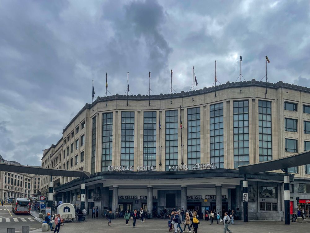 A wide view of Bruxelles-Central station on a cloudy day, with people walking across the plaza, buses passing on the street, and rows of flags lining the curved rooftop of the building.