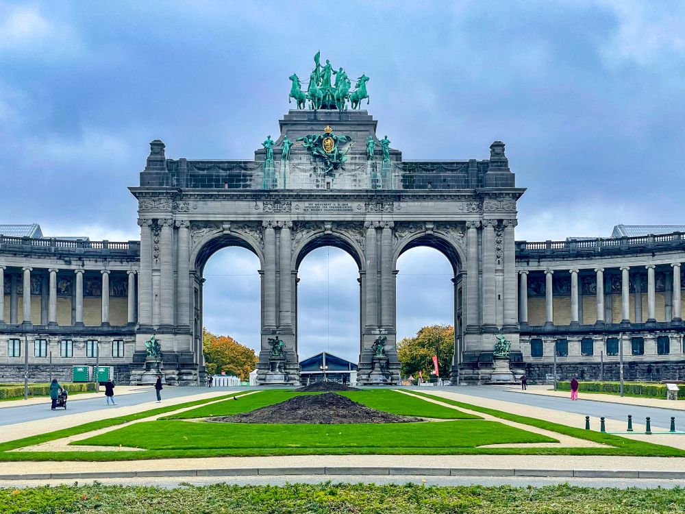 Photo of the Cinquantenaire Arch in Brussels, Belgium — a grand stone monument with three arches and a bronze sculpture on top, framed by green lawns and a cloudy sky with a few people walking nearby.