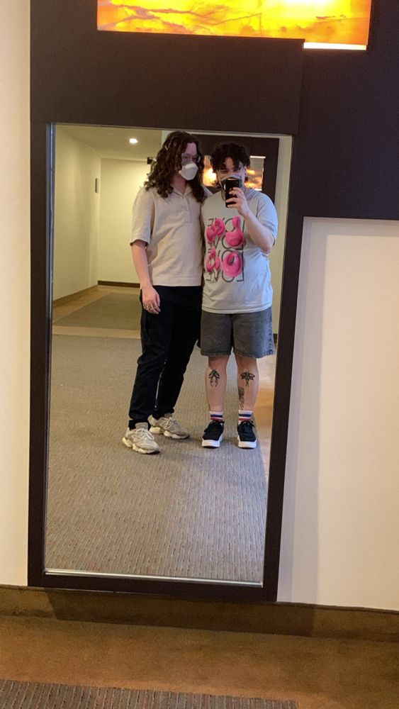 mirror selfie of edu and cb. cb has long dark curly hair, wearing glasses, a mask, a pale t-shirt and black pants. edu has short dark curly hair in a half ponytail, wearing a mask aswell, a blue tshirt with a design on it and grey pants. they're both embracing each other while edu is taking the picture 