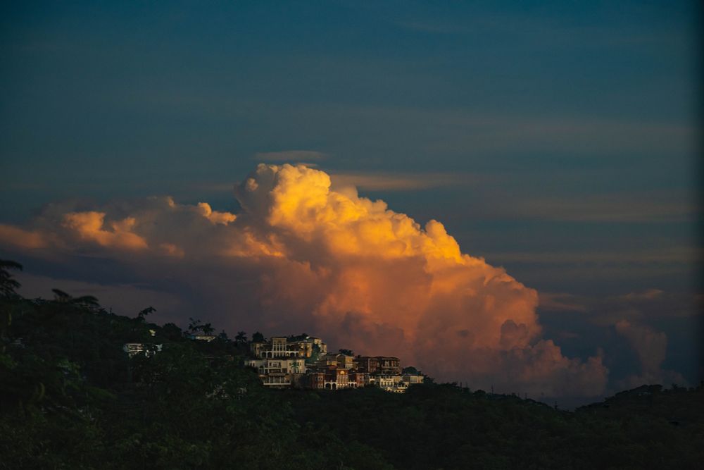 Sunset over Cruz Bay, St. John, USVI. Golden light catches a towering cloud formation above hillside villas, while the foreground remains in deep green shadow, creating a calm yet dramatic contrast between warmth and dusk.