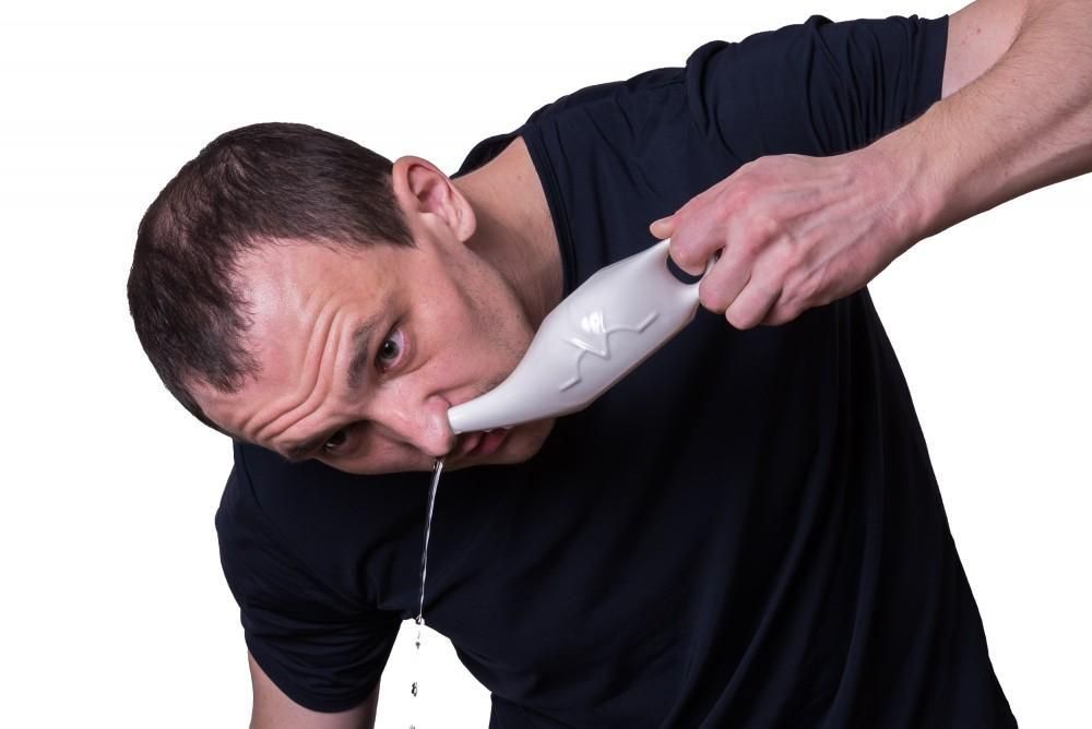 A man with thinning hair wearing a black t-shirt leaning forward with a neti pot jammed into one nostril; water is flowing out from the other.