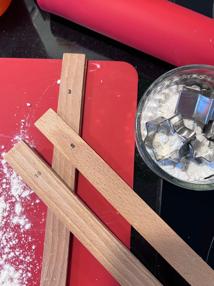 Teighölzer mit den Aufdrucken 3, 5 und 10 liegen auf einer roten Silikonmatte, daneben ein Glasschälchen mit Mehl und Ghetzliausstechern, oben ein rotes Silikonwallholz
