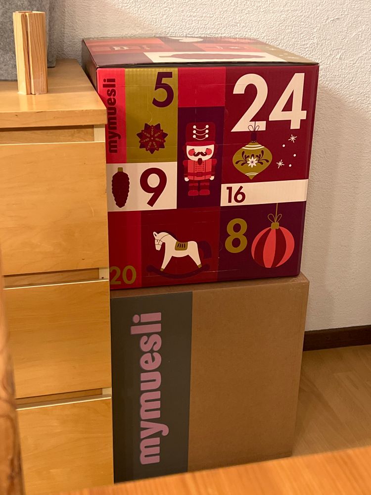 Ein MyMuesli Adventskalender Würfel auf der ebenso grossen Versandbox. Daneben eine Kommode, die fast gleichhoch ist wie die beiden Boxen. 