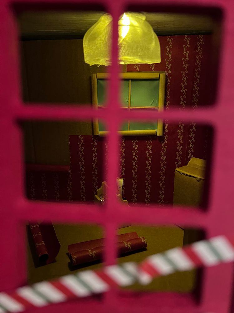 Blick durch die rote Wichteltür ins beleuchtete Wichtelhaus. Rechts steht eine Kiste, unter einem Fenster steht eine Werkzeugkiste, davor liegen Tapetenrollen. Die Wand ist noch nicht fertig tapeziert. 