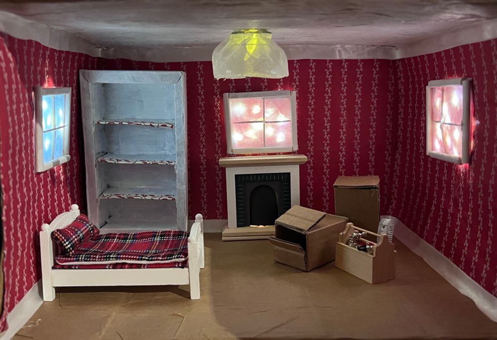 Gesamtansicht Wichtelhausinneres: links, rechts und in der Mitte je ein beleuchtetes Fenster, links steht ein leeres Regal, davor ein Bett, in der Mitte ein Kamin, rechts zwei Umzugskisten und ein Werkzeugkasten, an der Decke hängt eine Lampe, die Wände sind rot tapeziert, der Boden braun, die Decke weiss, oben und unten Bordüre/Leiste aus Textilband