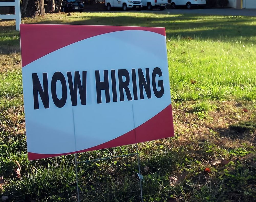 A red and white lawn sign that reads "Now Hiring" in black capital letters. 