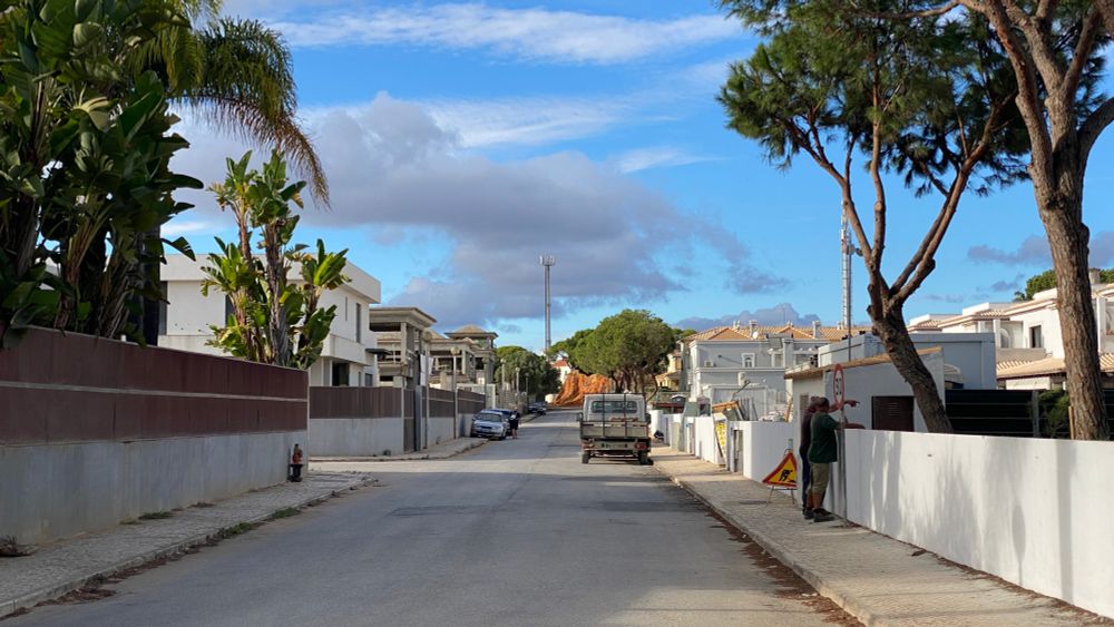 A street scene in ... well, that's the question!
What you can see, is palm trees, possibly bananas, white garden walls, a red and yellow work sign, an old pick up truck and some newly built apartment buildings. 
