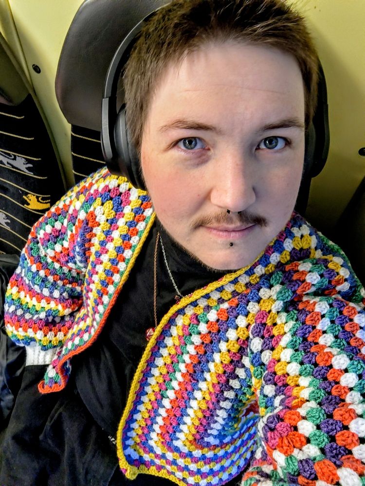 Ein*e Person sitzt in einem Sitz mit dunkler Kopfstütze, trägt große Kopfhörer und eine bunt gehäkelte, mehrfarbige Jacke aus kleinen Granny-Square-ähnlichen Mustern. Die Person hat kurzes Haar, einen dezenten Schnurrbart und ein Piercing unter der Unterlippe und schaut direkt in die Kamera.