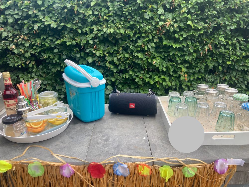 Eine improvisierte Cocktailbar mit Gläsern, in denen Limetten, Zitronen und Orangen in Scheiben geschnitten für Getränke bereit stehen. Dazu ein Glas Zucker , wiederverwendbare Strohhalme, ein Cocktail-Shaker und Grenadine -Sirup. 
Rechts daneben steht eine kleine Kühlbox, daneben eine schwarze JBL-Box und ein Tablett mit Gläsern.

An alle: Natürlich gibt es nur Saft-Cocktails, keinen Alkohol!