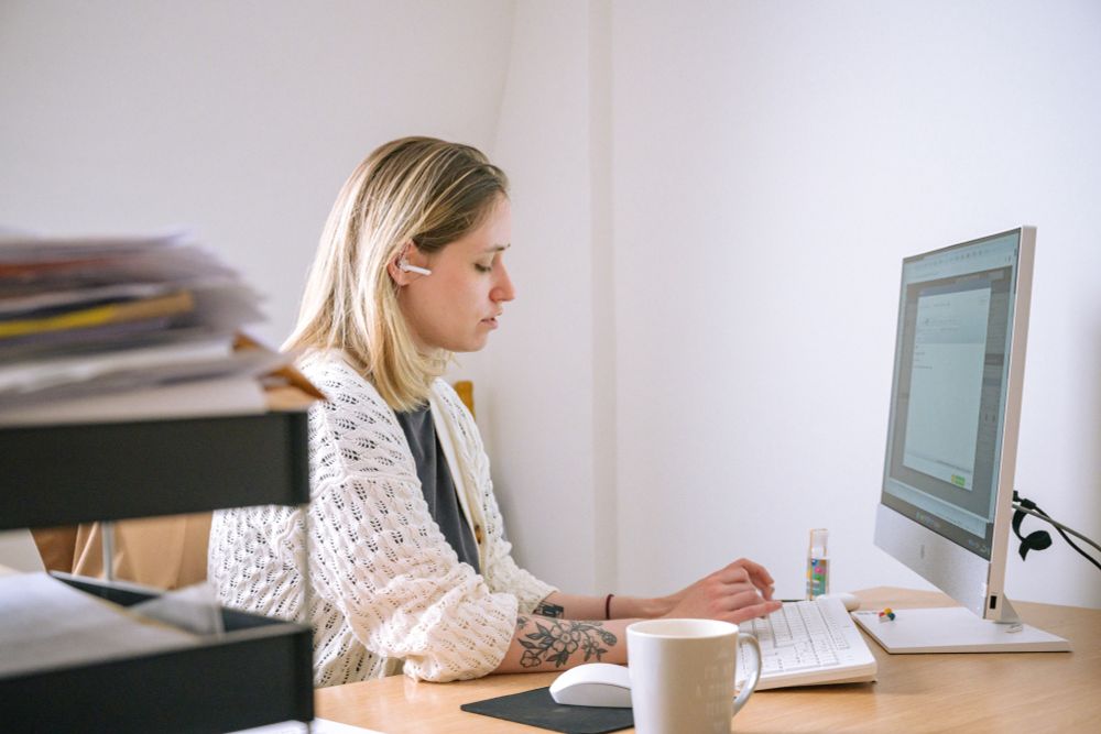 A person seen in side profile sat in front of a computer, typing on the keyboard. They are wearing a small headset and appear to be speaking to someone on the phone. In the foreground is a cup and an in-tray filled with paperwork.