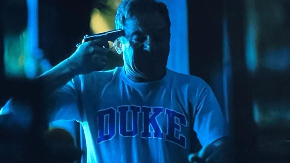 An image of Timothy Isaacs playing Timothy Ratliff in The White Lotus, with a gun to his head in a Duke t-shirt