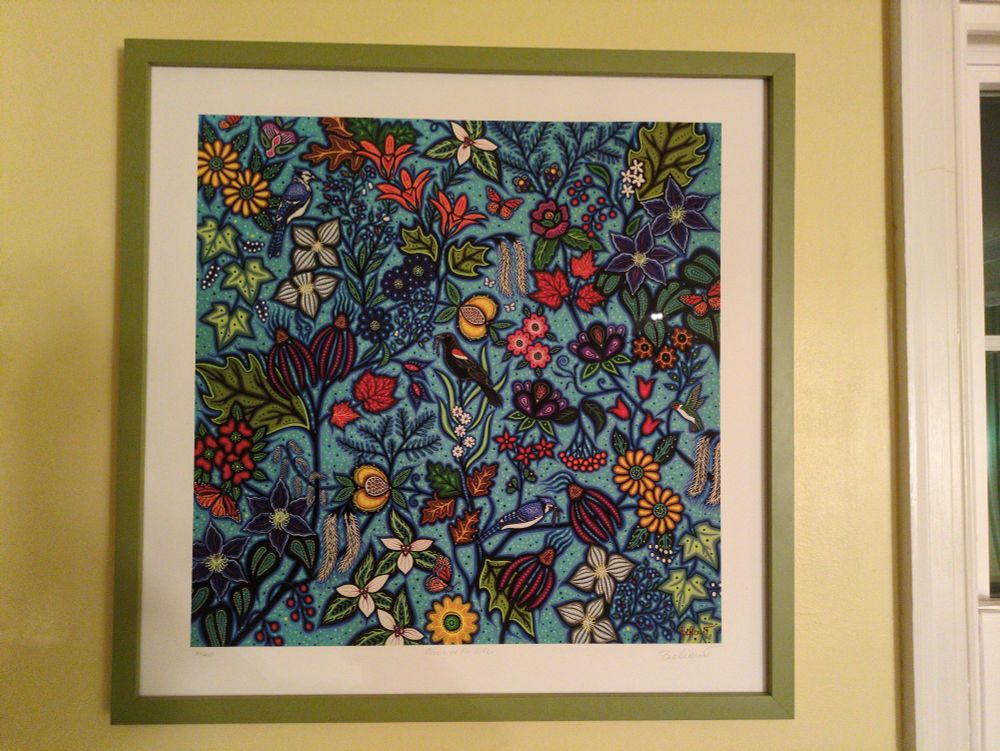 photo of a framed print by Christi  Belcourt depicting birds, butterflies, and plants on a blue background. It's really cool... I'm really sorry you can't see it  
