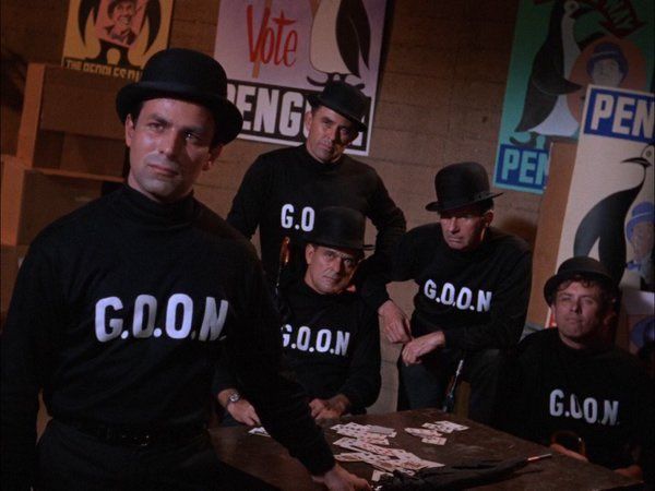 A screen grab from the 1960s batman series. A group of men wearing bowler hats and long sleeve black shirts with the word GOON written on them.