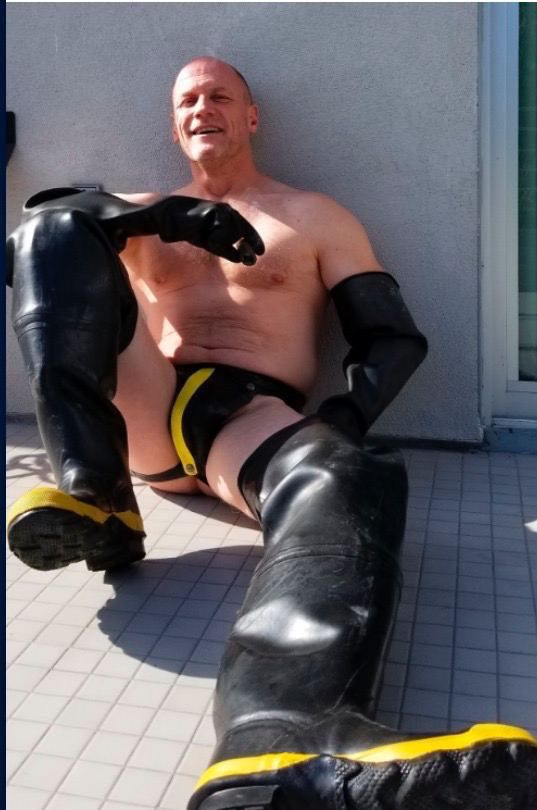 A photo of a bald man in a rubber jock, industrial rubber gloves and thigh high waders, sat on the ground casually and smiling 