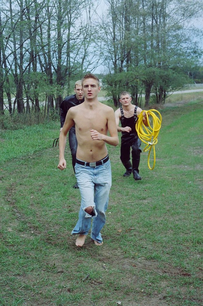 A photo of a shirtless man in jeans running across a field as two men dressed in latex overalls chase him.