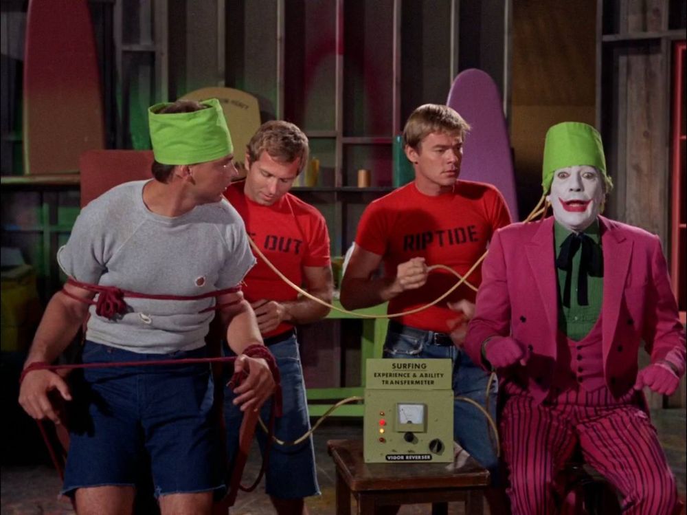 A screen grab from the 1960s batman series. Joker is in a chair next to a surfer connected by strange helmets. Behind them are two blonde musclemen wearing red shirts, one says Wipe Out, the other says Riptide.