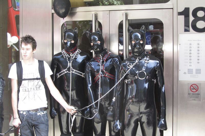A photo of 4 men on a subway train, one man is in a shirt shirt and he is holding leash to 3 identical rubber dogs all in matching catsuits, mitts and dog hoods.