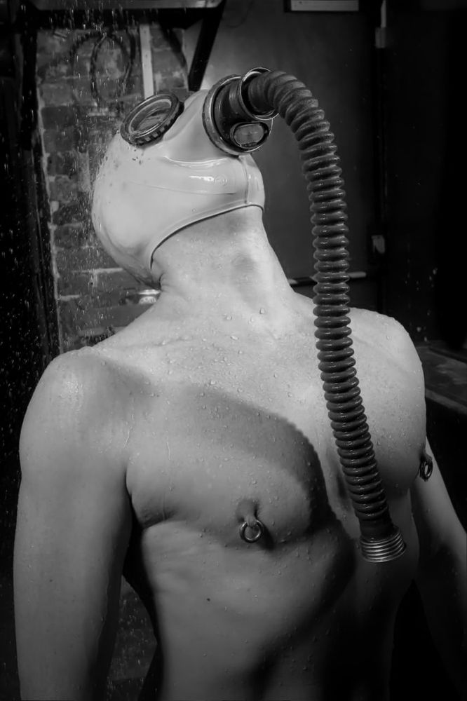 A black and white photo of a man leaning backwards dramatically whilst wearing a gasmask. He has large nipples that are both pierced with rings.