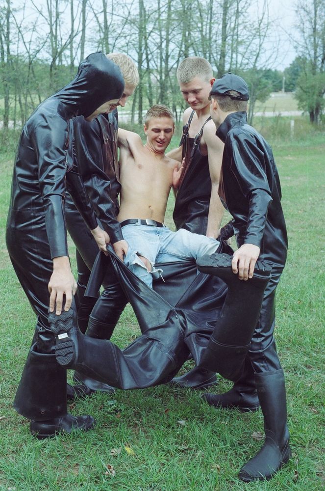 A photo of four rubber men lifting up the shirtless man and sliding him into a matching rubber suit.