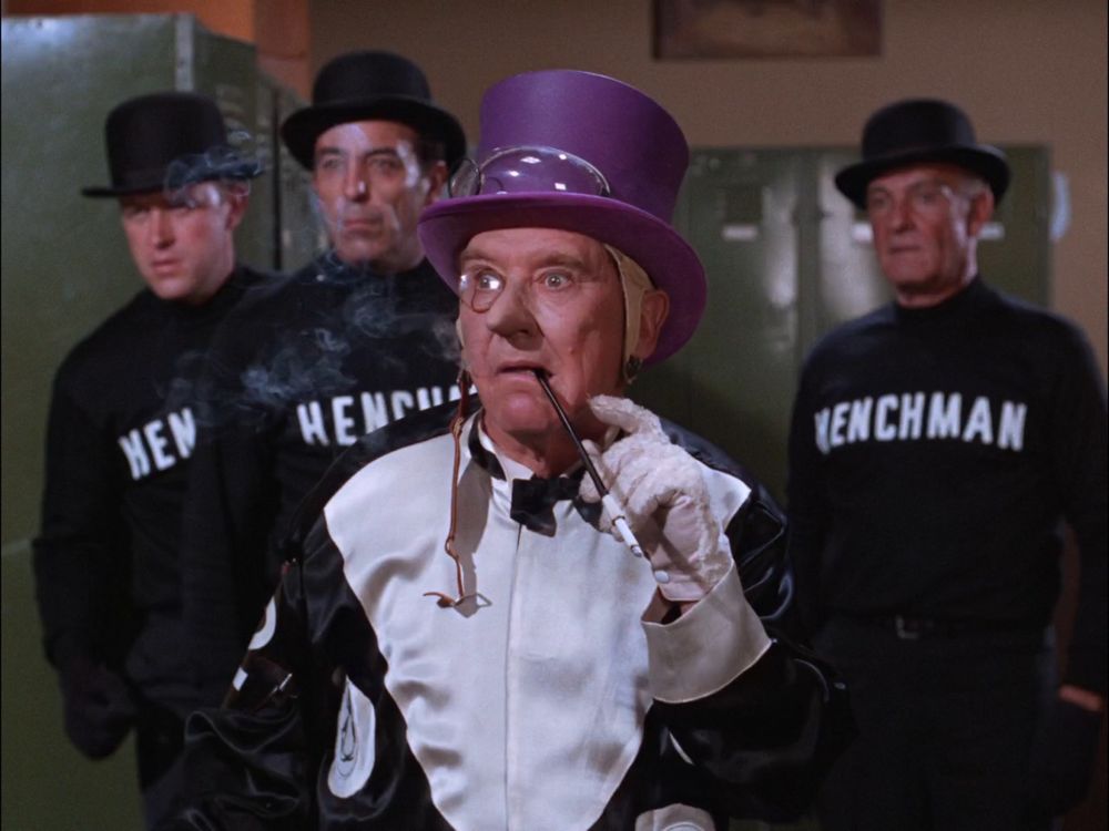 A screen grab from the 1960s batman series. A group of men wearing bowler hats and long sleeve black shirts with the word HENCHMAN written on them.