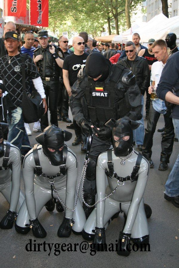 A photo of 3 identical rubber dogs wearing matching rubber suits and leather dogs hoods on their knees on a leash of a man in swat gear. Photographed at Folsom Berlin.