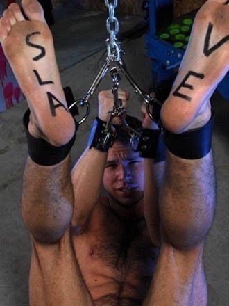 A photo of a man in bondage, he is naked with cuffs around his wrists and ankles and he is being hoisted upwards by these restraints, on the base of his feet someone has written the words SLAVE onto the soles of his feet.