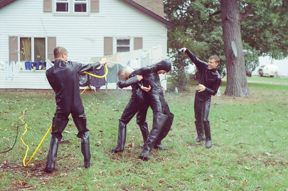 A photo of two men back to back, their arms linked, as two other rubber boys spray them with hoses in a garden.